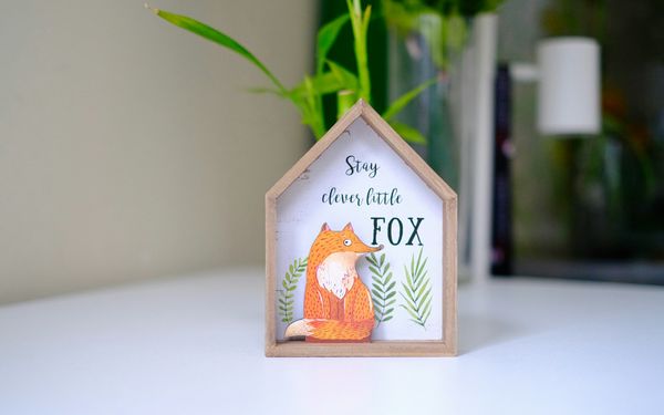 A small wooden house-shaped ornament on a desk, painted with a fox and the words Stay clever little FOX