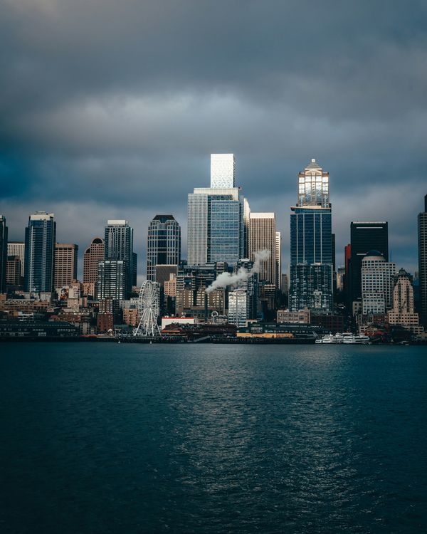 A city skyline of high-rise towers seen across dark water under heavy storm clouds